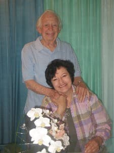 Frank Alper standing with Katharina Alper, his hands resting gently on her shoulders, a bouquet of white orchids before them.